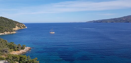 Blick auf felsige KĂĽsten vor Poros, umgeben vom blauen Meer und einem Segelboot
