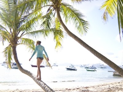 Mädchen balanciert auf einer schrägen Palme am Strand, im Hintergrund sind Yachten und Meer  Mädchen balanciert auf einer schrägen Palme am Strand, im Hintergrund sind Yachten und Meer